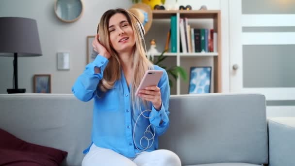 Retrato de joven linda chica joven atractiva en el acogedor fondo de la sala de estar escuchando música con auriculares. Mujer con blusa azul disfrutando de la música y bailando en casa . — Vídeos de Stock