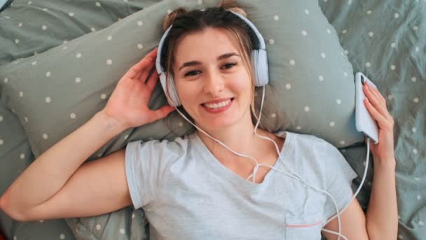Vue de dessus sur la jeune belle caucasienne souriante dans des écouteurs allongés sur le lit avec son smartphone et écoutant la musique tout en riant à la caméra. Ferme là. Vue d'en haut . — Video