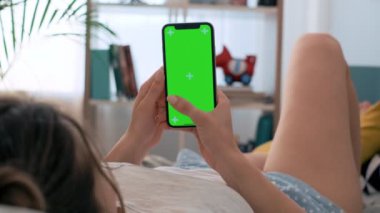 Sabah evde yatakta dinlenen ve yeşil ekranlı dikey akıllı telefona dokunan genç Beyaz kızın omzundan görünümü. Renk anahtarı.