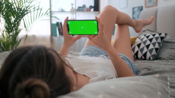 緑の画面と手の動きを追跡するスマートフォンで寝室のベッドに横たわっている若い白人女性の肩の上にビュー。クロマキー. — ストック動画