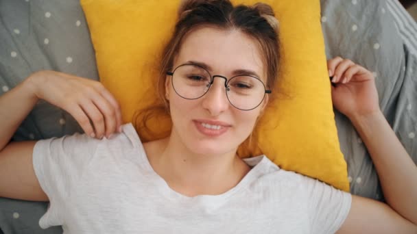 Prohlédněte si shora mladou veselou běloskavou ženu v brýlích, které leží na posteli, zívají a usmívají se na kameru. Zavři to. Portrét. Pohled shora. — Stock video