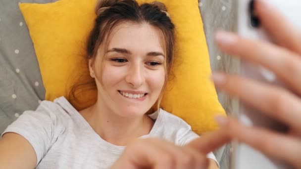 Vue du dessus de la jeune caucasienne souriante jolie fille couchée sur le lit et allumant la webcam sur le smartphone comme ayant vidéochat. Ferme là. Vue d'en haut . — Video