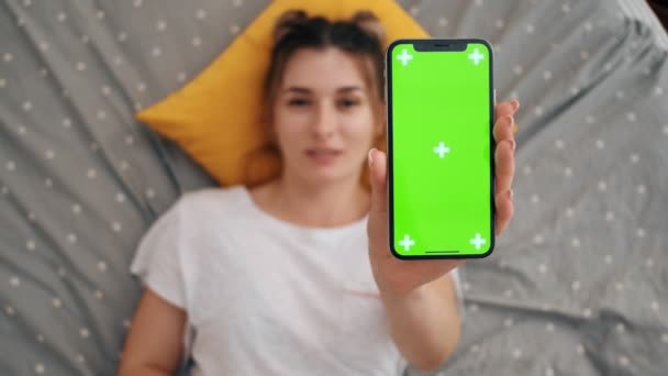 Nahaufnahme des schwarzen Smartphones mit grünem Bildschirm und Tracking-Bewegung, das eine junge hübsche Frau auf dem Bett liegend der Kamera vorführt. Ansicht von oben. Blick von oben. — Stockvideo