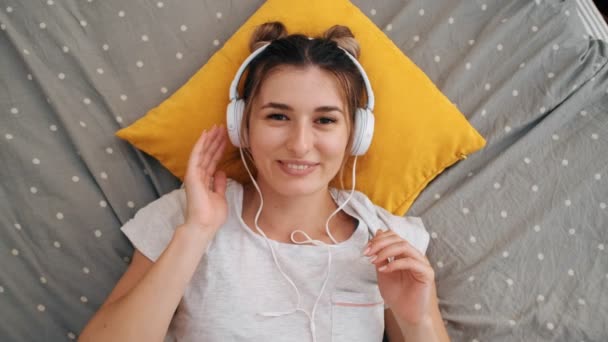 Vue du dessus du portrait de la jeune fille heureuse caucasienne charmante dans des écouteurs couchés sur le lit et souriant à la caméra. Ferme là. Vue d'en haut . — Video