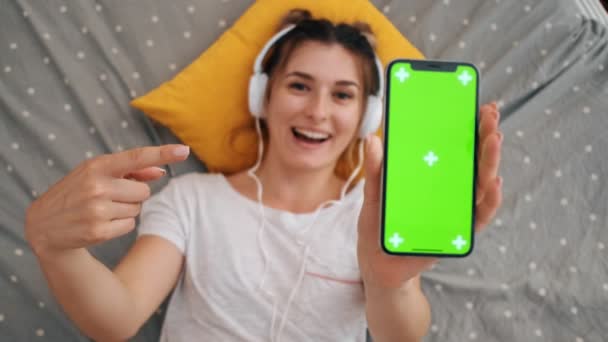 Close up of the black smartphone with green screen and tracking motion which woman demonstrating to the camera and pointing with a finger at it while lying on the bed in the headphones. — Stock Video