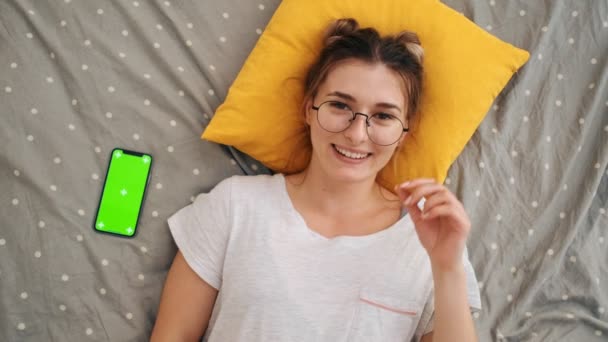 Vista de cima na menina jovem de óculos deitada na cama, sorrindo alegremente para a câmera e apontando com um dedo para o smartphone com tela verde e movimento de rastreamento. Chave Chroma . — Vídeo de Stock