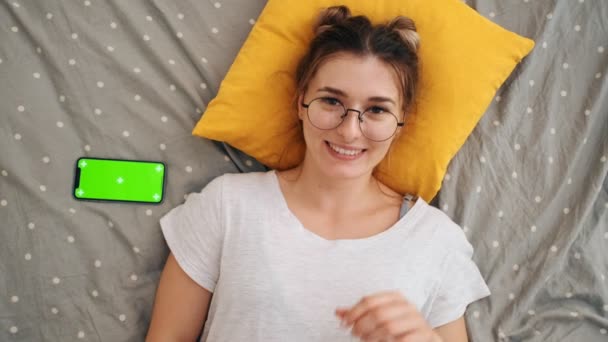 Visão superior sobre a jovem menina encantadora em óculos deitados na cama, sorrindo alegremente para a câmera e apontando com um dedo para o smartphone com tela verde e movimento de rastreamento. Chave Chroma . — Vídeo de Stock
