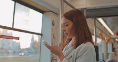 Akıllı telefon tarama internet ve arkadaşlarınızla sohbet kullanarak toplu taşıma çekici gülümseyen kız portre. Tramvay üzerinde seyahat ederken sevimli kadın, el parmaklık tutar. Şehir, kentsel arka plan.