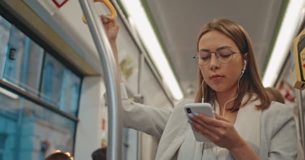 Porträt des netten brünetten Mädchens mit Brille und Kopfhörer hält den Handlauf, hört Musik und surft mit dem Handy in öffentlichen Verkehrsmitteln. — Stockvideo