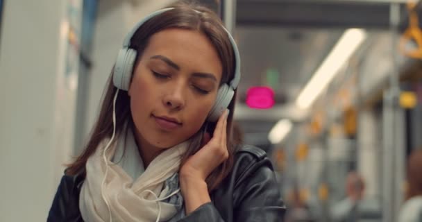 Feliz chica sonriente en auriculares escuchando música y navegando en el teléfono móvil en el transporte público por la noche. Ciudad luces fondo . — Vídeos de Stock