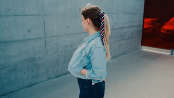 Retrato menina branca bonita e elegante na jaqueta jeans de pé na rua da cidade perto da parede, voltando-se para a câmera e sorrindo . — Vídeo de Stock