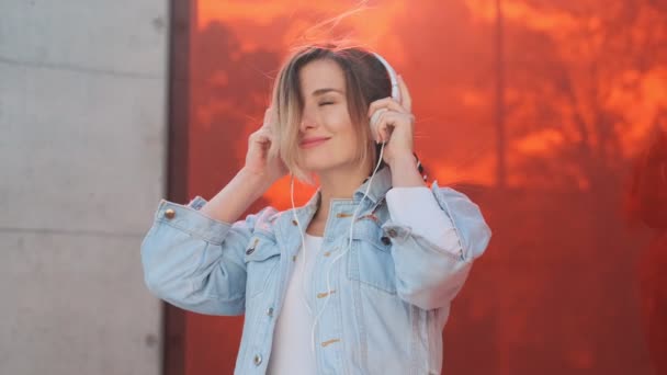 Cheerful young Caucasian pretty stylish woman smiling and having fun while taking on headphones and listening to the music on the street of the city. — Stock Video