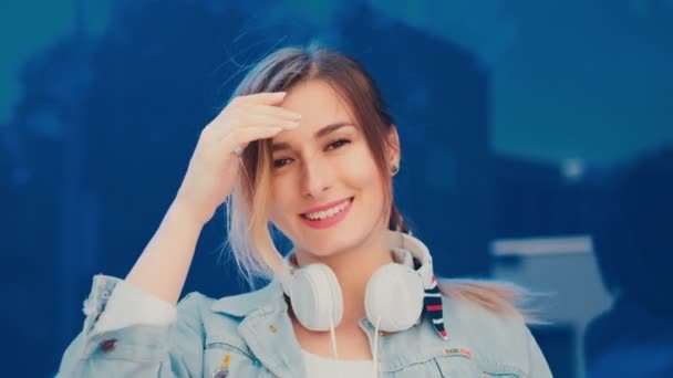 Retrato de la encantadora y feliz joven y elegante chica con unos grandes auriculares blancos en el cuello mirando a la cámara y riendo alegremente mientras está de pie al aire libre sobre el fondo de cristal azul . — Vídeo de stock