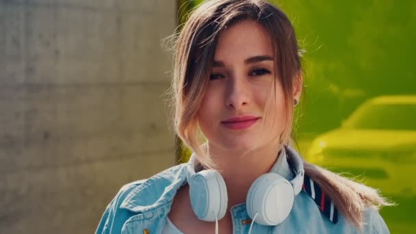 Porträt der schönen und glücklichen kaukasischen jungen Frau mit einem großen weißen Kopfhörer auf dem Hals, die fröhlich in die Kamera draußen im Wind lächelt. Nahaufnahme. — Stockvideo