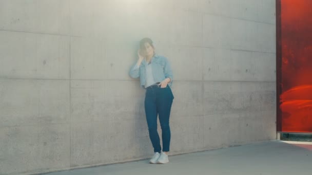 Caucásico joven mujer elegante y bonita en los pantalones vaqueros, chaqueta y grandes auriculares blancos escuchando la música mientras se apoya en la pared en la calle . — Vídeo de stock