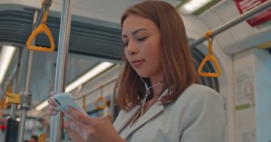 Kulaklık sevimli esmer kız yakın portre toplu taşıma cep telefonu tarama tırabzan tutar. Akıllı telefonu kullanan kadın kulaklıkla müzik dinlemesohbeti yapıyor