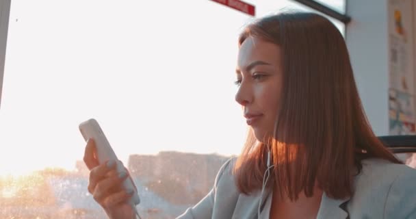 Porträtt av söt leende flicka i hörlurar lyssnar på musik och surfar på smartphone i kollektivtrafiken. Lycklig kvinna tittar ut ur ett tåg fönster, tänker på något under sin dagliga pendla — Stockvideo