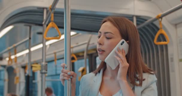 可爱的女孩的肖像拿着扶手，在公共交通工具上用手机说话。快乐有吸引力的女人站在城市电车在她的日常通勤和智能手机说话. — 图库视频影像
