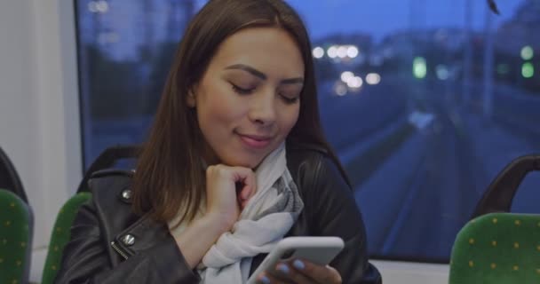 Ritratto di giovane ragazza attraente con smartphone durante la corsa in tram in serata in città. Donna nei trasporti pubblici utilizzando smartphone navigazione internet e chattare con gli amici. Luci notturne . — Video Stock