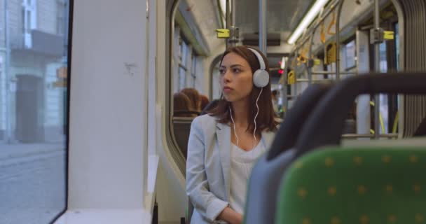 Retrato de menina séria em fones de ouvido. Ela usa smartphone para ouvir música. Mulher atraente sentada perto da janela no bonde da cidade durante seu trajeto diário e ouvir música . — Vídeo de Stock