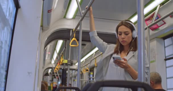 Retrato de una chica sonriente que usa audífonos sostiene la barandilla y usa un teléfono inteligente, escuchando música. Mujeres atractivas navegando en el teléfono celular en el transporte público. Ciudad, transporte, conmutar, urbano . — Vídeo de stock