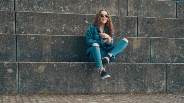 Портрет молодой кавказской стильной хипстерки в джинсовой одежде и темных солнцезащитных очках, сидящей на больших обкуренных ступеньках и скрещивающей ноги . — стоковое видео