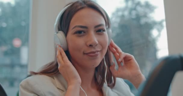 Cierre de la guapa joven caucásica en los grandes audífonos blancos sonriendo a la cámara y escuchando la música mientras se va en el tranvía. Retrato. — Vídeo de stock