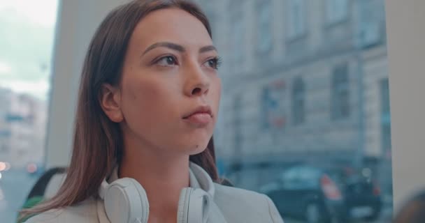 トラムやバスのどこかに行く間、彼女の首に大きな白いヘッドフォンと窓を見ている若い白人の美しい女性。閉じろ!. — ストック動画