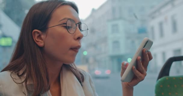 La joven caucásica guapa en gafas tapping y texting en el teléfono inteligente mientras se sienta en el tranvía o autobús y regresa a casa. Cierre. — Vídeos de Stock