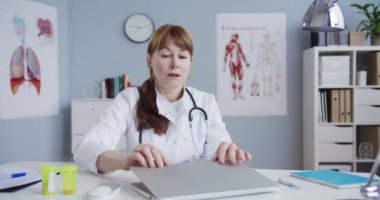 Beyaz kadın doktor, dolapta otururken bilgisayarı kapatıyor. Hastane ofisinde dizüstü bilgisayarla çalıştıktan sonra yorgun düşmüş güzel bir doktor. İlaç ve sağlık hizmetleri. Tıbbi konsept.