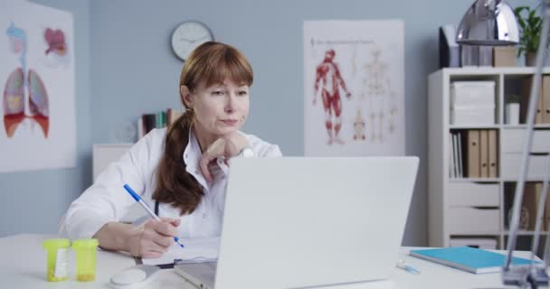 흰 가운을 입은 아름다운 여성 의사가 캐 비넷에 앉아 노트북으로 온라인상에서 상담을 받고 있습니다. 백인 의사는 의학적 서류를 작성하는 동안 웹캠을 통해 환자와 상담 한다. 건강 관리 개념 — 비디오