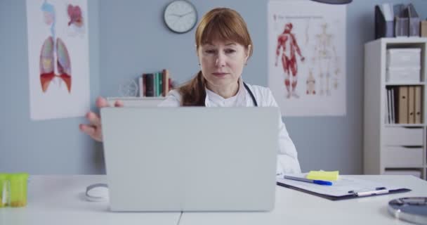 Uma médica bonita de vestido branco a trabalhar no computador na secretária do armário. Bela médica branca abrindo laptop enquanto estava sentada no consultório do hospital. Médico digitando no laptop . — Vídeo de Stock