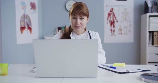 Mooie vrouwelijke arts typen op laptop in witte jurk aan bureau in kabinet. Mooie blanke vrouwelijke arts op zoek naar internet op de computer in het ziekenhuis kantoor. Gezondheidszorgconcept. — Stockvideo