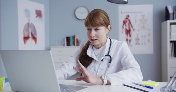キャビネットのテーブルに座って白いガウンを着た女性はかなり白人医師。医師はノートパソコンでビデオ撮影し、ウェブカメラを介して患者に相談します。オンラインでビデオチャットや相談を持つ医療. — ストック動画