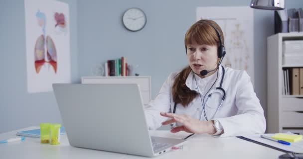 Jolie femme médecin caucasienne dans casque assis à table dans le cabinet et vidéochat sur ordinateur portable. Magnifique médecin consultant patient via webcam. Médecin ayant une consultation en ligne. Concept de santé — Video