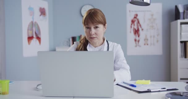 Dolabın masasında otururken bilgisayarda yazan güzel beyaz kadın doktor. Güzel mutlu doktor bilgisayarını kapatıp hastane ofisindeki kameraya gülümsüyor. Tıp konsepti. — Stok video