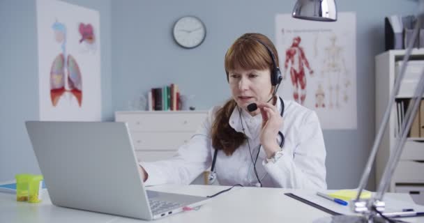 Beautiful doctor consulting patient via webcam. Hezká běloška ve sluchátkách sedí u stolu ve skříňce a natáčí na notebooku. Lékařská konzultace online. Koncept zdravotní péče — Stock video