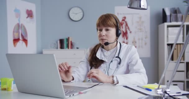 Beautiful female doctor in white gown consulting patient via webcam and showing medication. Pretty female Caucasian physician in headset sitting at table in cabinet and videochatting on laptop. — Stock Video