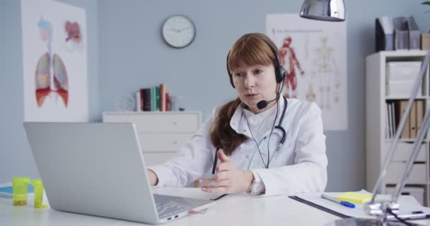 Güzel, beyaz kulaklıklı bir kadın doktor kabinede masada oturuyor ve dizüstü bilgisayarında online danışmanlık yapıyor. Güzel Doktor tedaviyi ve tedaviyi web kamerasıyla açıklıyor. Sağlık kavramı — Stok video