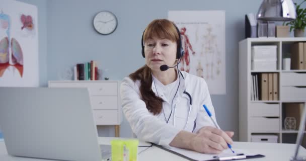 Bela médica caucasiana no fone de ouvido sentado à mesa no armário e escrevendo os sintomas. Doutor videochat no computador portátil e paciente consultoria via webcam. Consulta online . — Vídeo de Stock