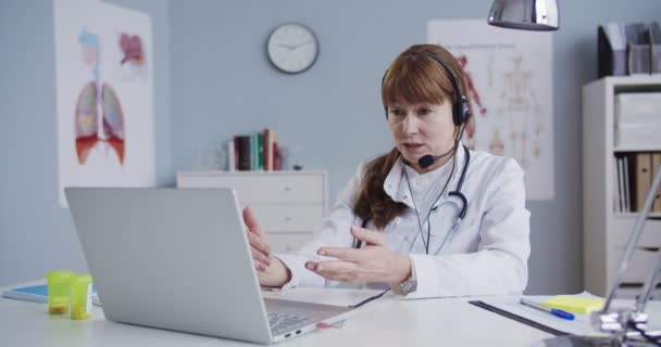 一位漂亮的女医生坐在橱柜里的桌子旁，她戴着耳机，在笔记本电脑上有在线咨询。美丽的医生演示并解释如何通过网络摄像头使用温度计。医学概念 — 图库视频影像