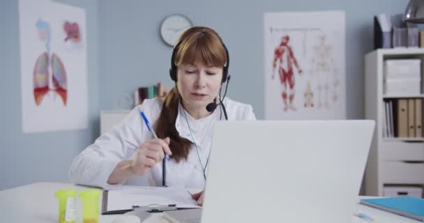 Médico caucásico que tiene videochat en el ordenador portátil y consultar al paciente a través de la webcam mientras llena el formulario médico. Hermosa mujer médico en auriculares sentado en el gabinete y tener consulta en línea . — Vídeo de stock