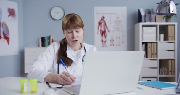 Beyaz cüppeli güzel beyaz kadın doktor, kabinede oturuyor ve laptopta tıbbi formları kontrol ederken online danışmanlık yapıyor. Web kamerasıyla doktora danışıyorum. Sağlık hizmetleri kavramı — Stok video