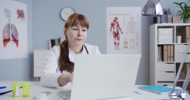 Güzel beyaz kadın doktor dolapta otururken bilgisayarda yazı yazıyor. Hastane ofisinde dizüstü bilgisayarla çalışan oldukça yorgun bir doktor. İlaç ve sağlık hizmetleri. Sağlık kavramı. — Stok video
