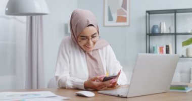 Geleneksel başörtüsü ve şık gözlüklü genç Müslüman kız bilgisayarla proje üzerinde çalışırken ve gülümserken akıllı telefonunu kullanıyor. Uzaktan çalışma, ev ofis konsepti.
