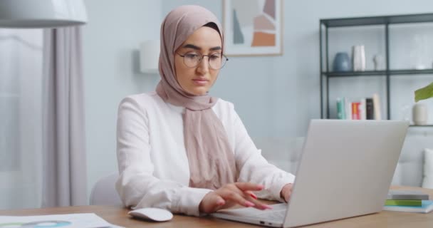 Joven hermosa mujer musulmana en gafas terminan de trabajar en la computadora. Chica segura y satisfecha mirando desde la pantalla del ordenador portátil a la cámara. Negocios exitosos, trabajo remoto, concepto de oficina en casa . — Vídeo de stock