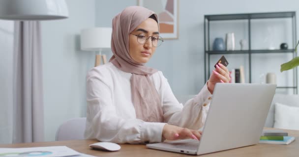 Plano medio de hermosa chica musulmana asiática en gafas elegantes compras en línea celebración de la tarjeta de crédito, utilizando el ordenador portátil en casa. Transacciones fáciles, banca en línea, transacciones y concepto de estilo de vida moderno . — Vídeo de stock