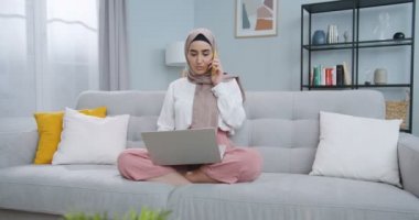 Orta plan, tesettürlü güzel müslüman bir kızın evde cep telefonuyla konuşup bilgisayar kullanırken arkadaşlarıyla haber konuşması. İnsan kavramı, iletişim ve ev kavramı.
