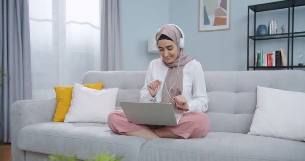 Střední plán drzé krásné muslimky v hidžábu poslech hudby v bílých sluchátkách a tanec při posezení na pohovce v moderním stylovém obývacím pokoji. Dívka se baví při pobytu doma. — Stock video