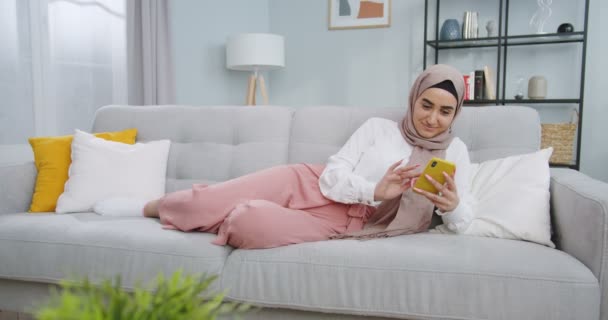 Средний план мусульманской женщины со смартфоном, смотрящей на экран мобильного телефона смеясь, наслаждаясь, используя мобильные приложения, веселясь, играя в игры чаты в социальных сетях лежа на диване дома . — стоковое видео